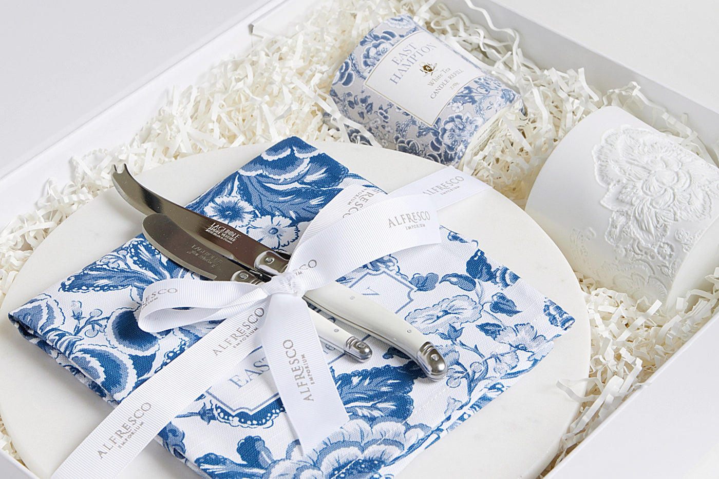A corporate Gift that contains a round marble cheese board, pate knife, cheese knife, blue and white tea towel, matching East Hamptons White Tea Scent candle refill and co-ordinating bisque candle vessel.