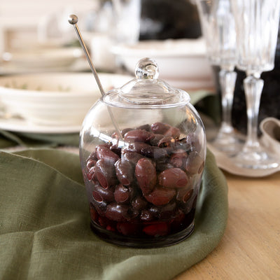 Glass Olive Jar With Metal Spoon Styled On Table With Olives