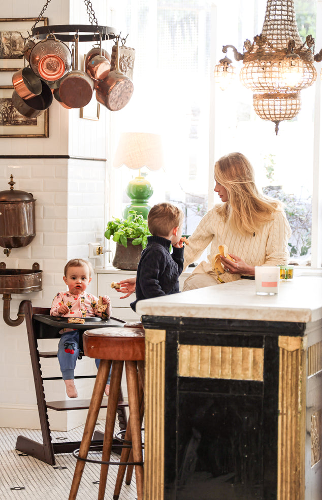A woman and two children share a meal at a rustic kitchen island, surrounded by hanging copper pots, a chandelier, and a bright window in a farmhouse-style kitchen.