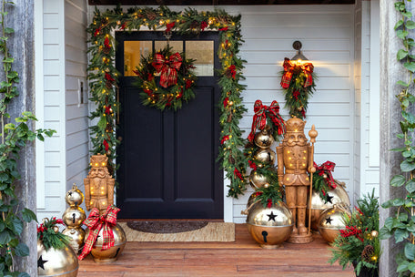 A festive holiday front porch decorated with green garlands, red bows, a wreath on the black door, gold nutcrackers, and large shiny gold ornaments.