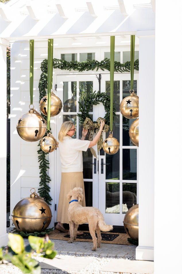 Laundry Door Decorated with Green Garland and Wreath and Oversized Gold Bells Hanging from Pergola Woman Tying Bow and Dog Watching