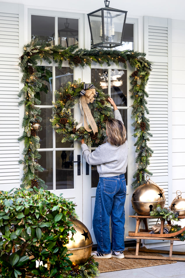 Woman Hanging blue and green wreath on Christmas Door styled with Garland and Oversized Gold Bells out front