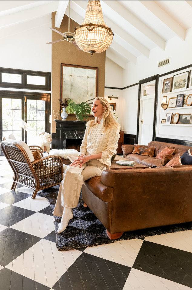 A woman sits on a brown leather sofa in a bright, stylish living room with a black and white checkered floor, a large woven chandelier, and a fireplace in the background.
