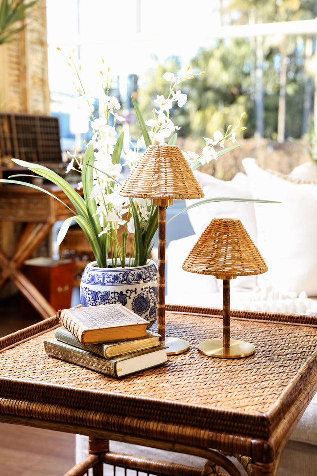 A styled interior scene showing a woven rattan coffee table with stacked books, a blue-and-white ceramic vase of white flowers, and two small Rattan cordless table lamps in a sunlit room.