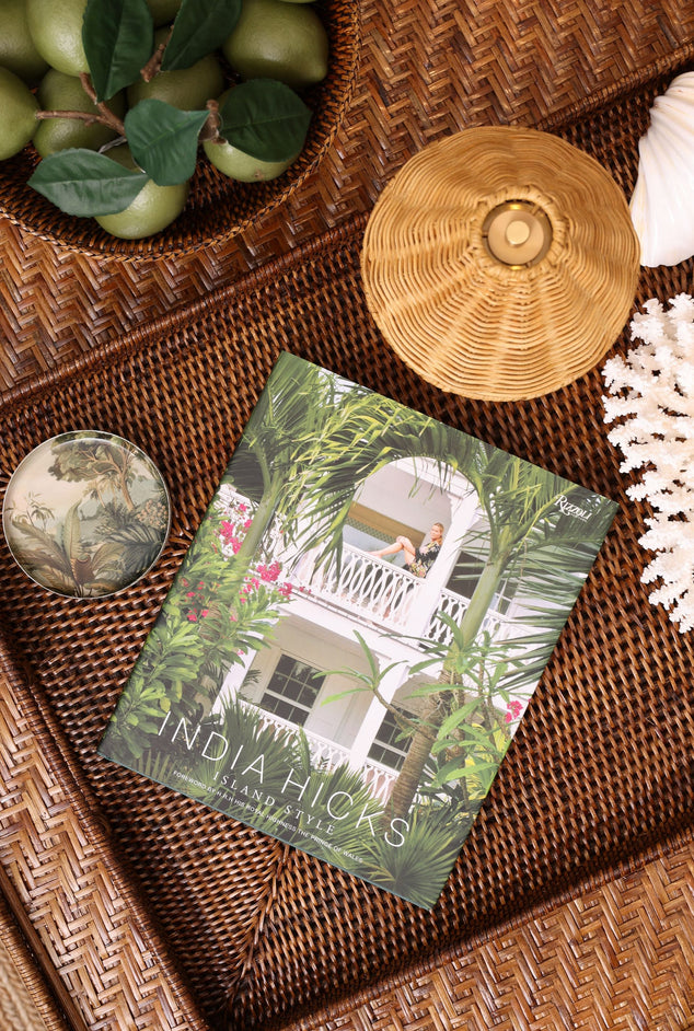 A flat-lay photograph of a book titled 'INDIA HICKS' resting on a brown woven rattan tray, surrounded by decorative props including green limes, a woven hat, a shell, and coral.