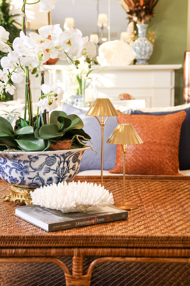 A styled interior scene with a wicker coffee table holding a blue-and-white ceramic planter with white orchids, a white coral-like decorative piece, a book, and two small gold table lamps. A sofa with blue and rust-orange pillows is blurred in the background.
