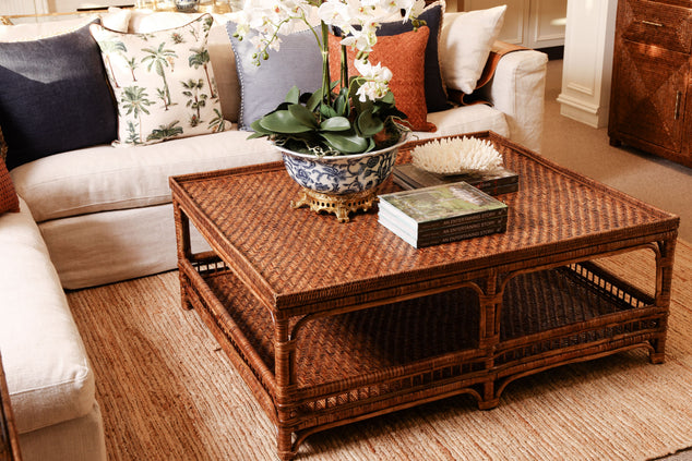 A rectangular rattan coffee table with a lower shelf, styled in a living room with a cream sofa, decorative pillows, and a potted orchid on top.