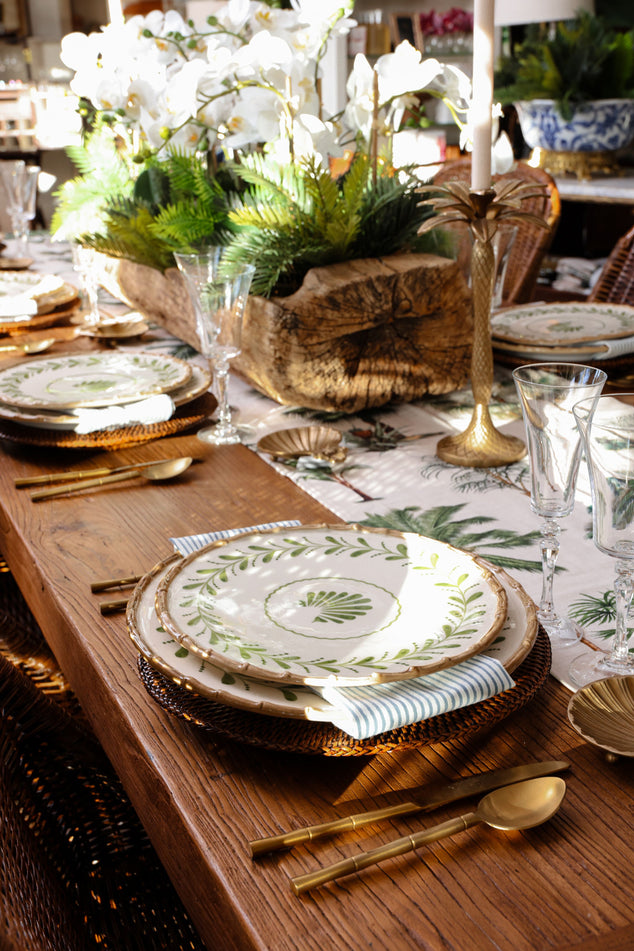 A styled dining table setting with stacked white plates featuring green botanical leaf patterns, gold flatware, woven placemats, and a rustic wood centerpiece box filled with white flowers and greenery.