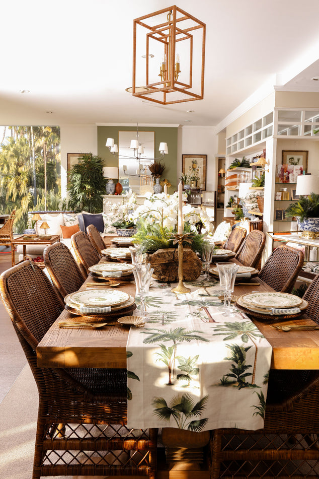 A dining room set with a rectangular wooden table, wicker dining chairs, a white table runner with a green palm leaf pattern, and a large geometric copper-toned pendant light hanging above.