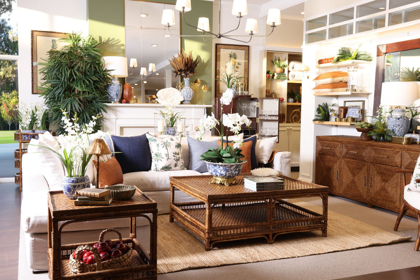 A styled living room interior featuring a cream sofa with navy and floral throw pillows, a rectangular woven rattan coffee table, a matching side table, a large beige area rug, and warm wood cabinetry in the background.