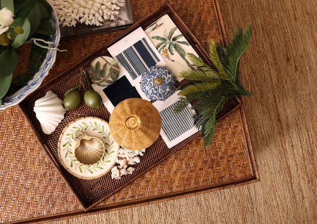 A styled flat-lay of home decor and textiles on a woven tray, including patterned fabric swatches, a small blue-and-white ceramic pot, seashells, green olives, a woven basket lid, and tropical leaves on a woven mat.
