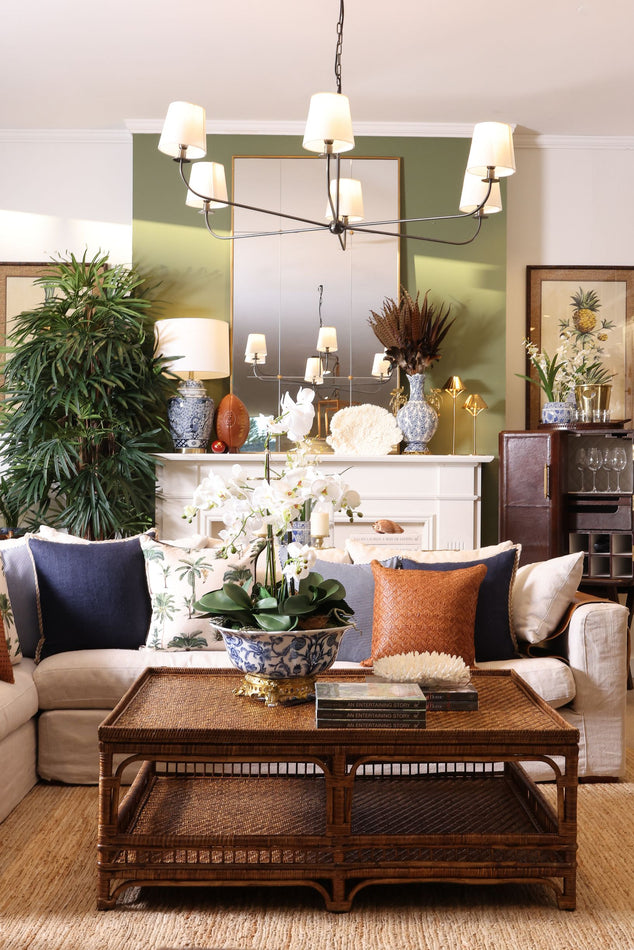 A styled living room interior featuring a dark brown wooden coffee table with a lower woven shelf, set in front of a beige sectional sofa with navy and rust accent pillows. The room has a green accent wall, a white console table with lamps, and a multi-light chandelier overhead.
