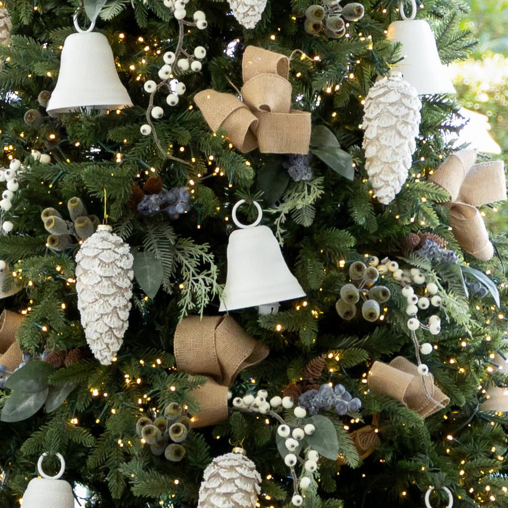 A close-up of a decorated Christmas tree with white bell-shaped ornaments, large textured pinecone ornaments, burlap ribbon bows, green foliage, and warm string lights.