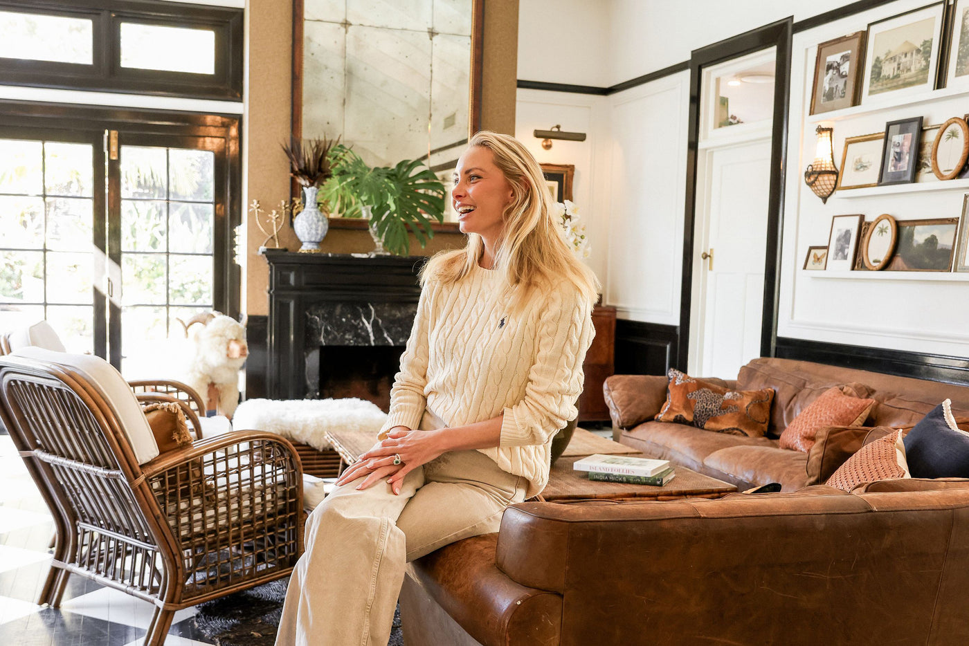Fiona sits smiling on a brown leather sofa in a cozy, traditional living room. She is wearing a cream cable-knit sweater and matching cream trousers.