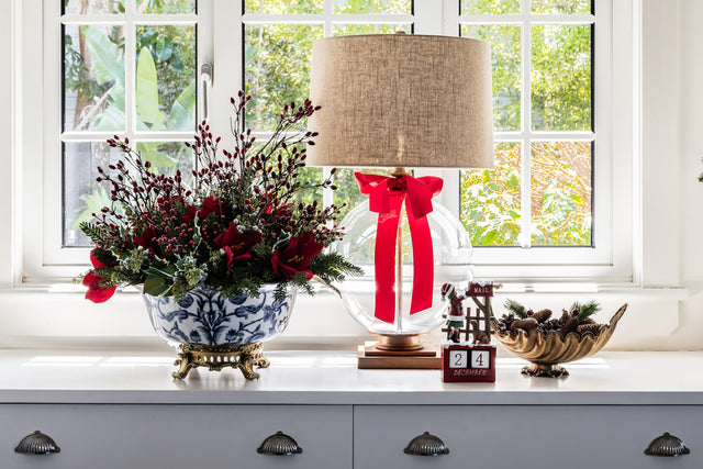 A blue and white ceramic bowl filled with red berry spays, a lamp with red bow and gold dish with pinecones.