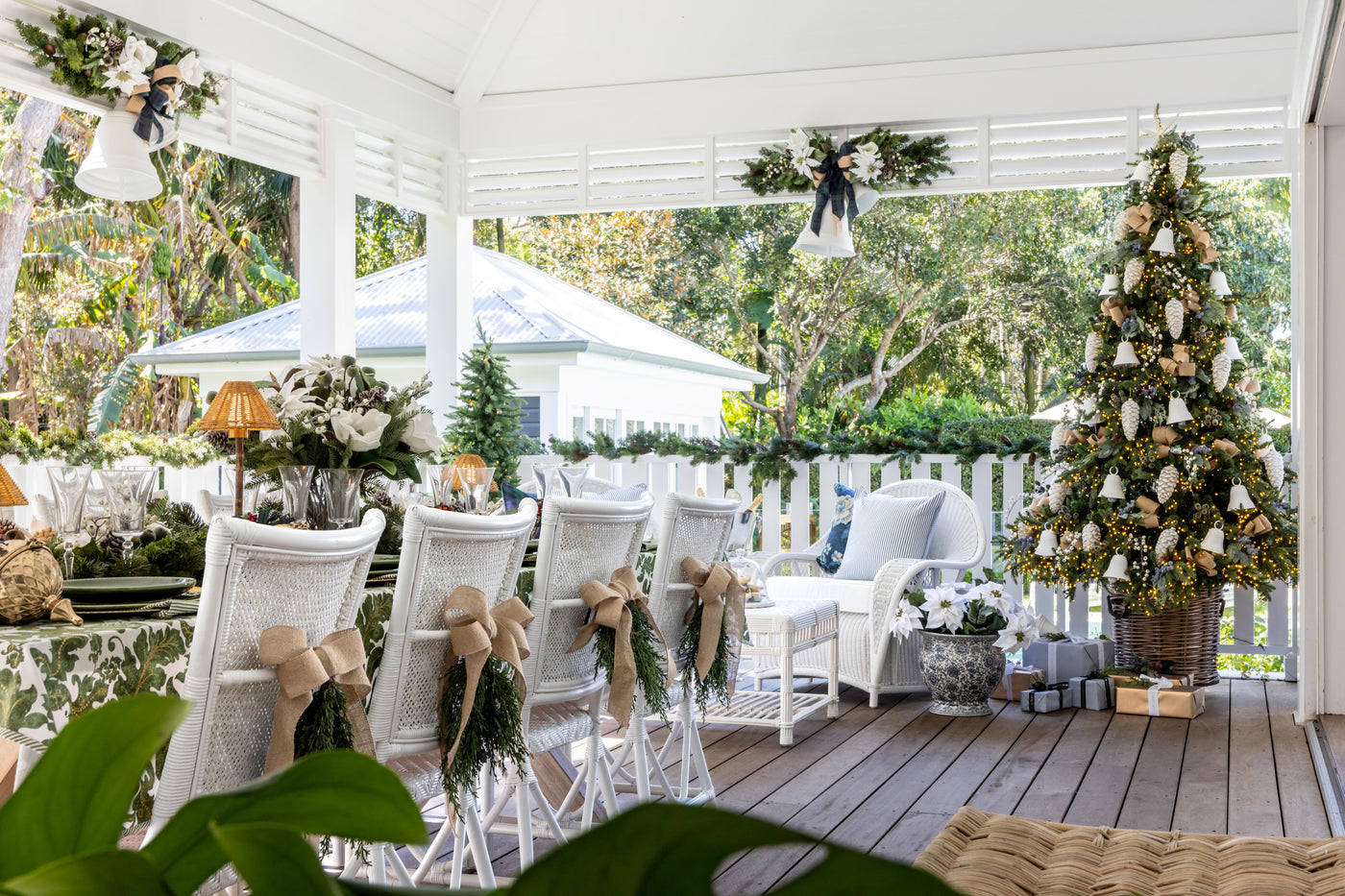 A decorated outdoor porch with white wicker dining chairs adorned with bows and green garlands, a white armchair with a blue cushion, a Christmas tree with white ornaments, and a white fence in the background.