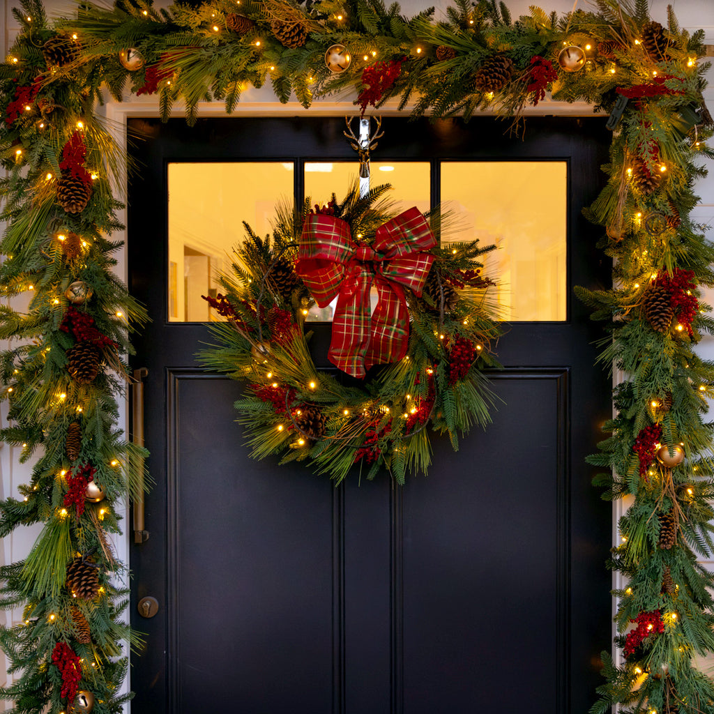 Decorative red berry Christmas wreath with a red bow on a black door, surrounded by garland and lights.