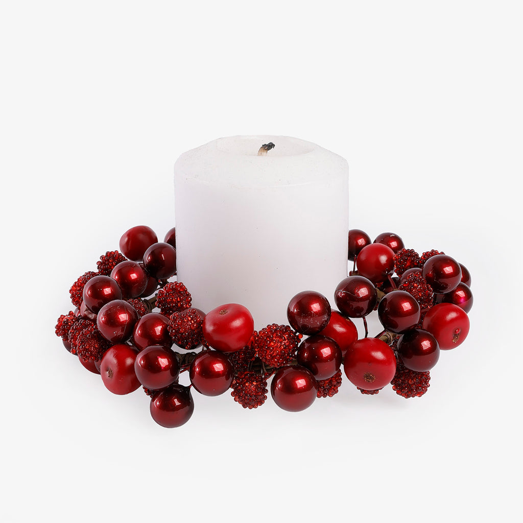 A white pillar candle surrounded by a decorative wreath of glossy red berries and textured red foliage on a clean white background.