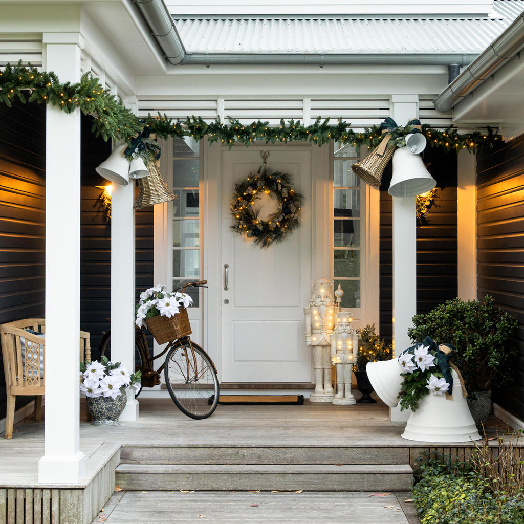 A beautifully decorated front porch with a white door, white columns, a green garland, a wreath, a bicycle with flowers in the basket, and decorative bells and planters.