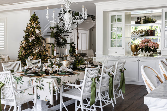 White rattan chairs around a dining table covered with a palm tree tablecloth. A Christmas tree is in the background with palm tree baubles and orchids.