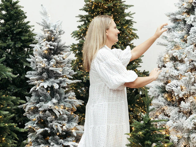 A lady in a white dress surrounded by evergreen and snow artificial Christmas trees.