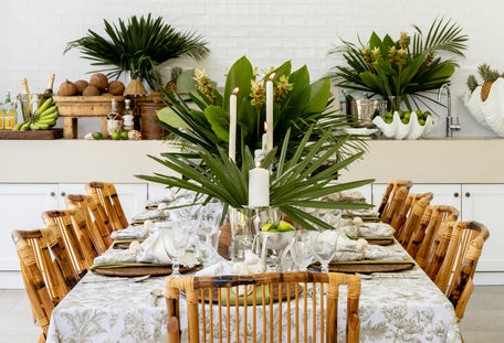 A styled dining table setup in a bright outdoor area, featuring a white and beige toile patterned tablecloth.