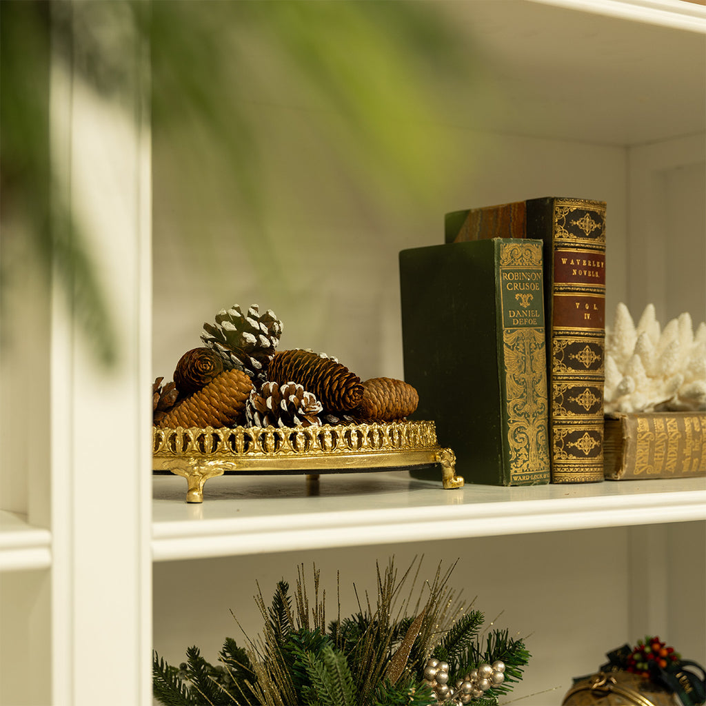 A gold mirror tray full of pinecones on a white bookshelf.
