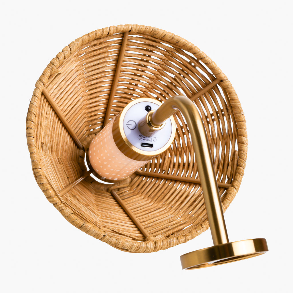 A modern wall lamp with a large circular woven rattan shade, a curved brass metal arm, and a peach-colored cylindrical light bulb holder, shown against a plain white background.