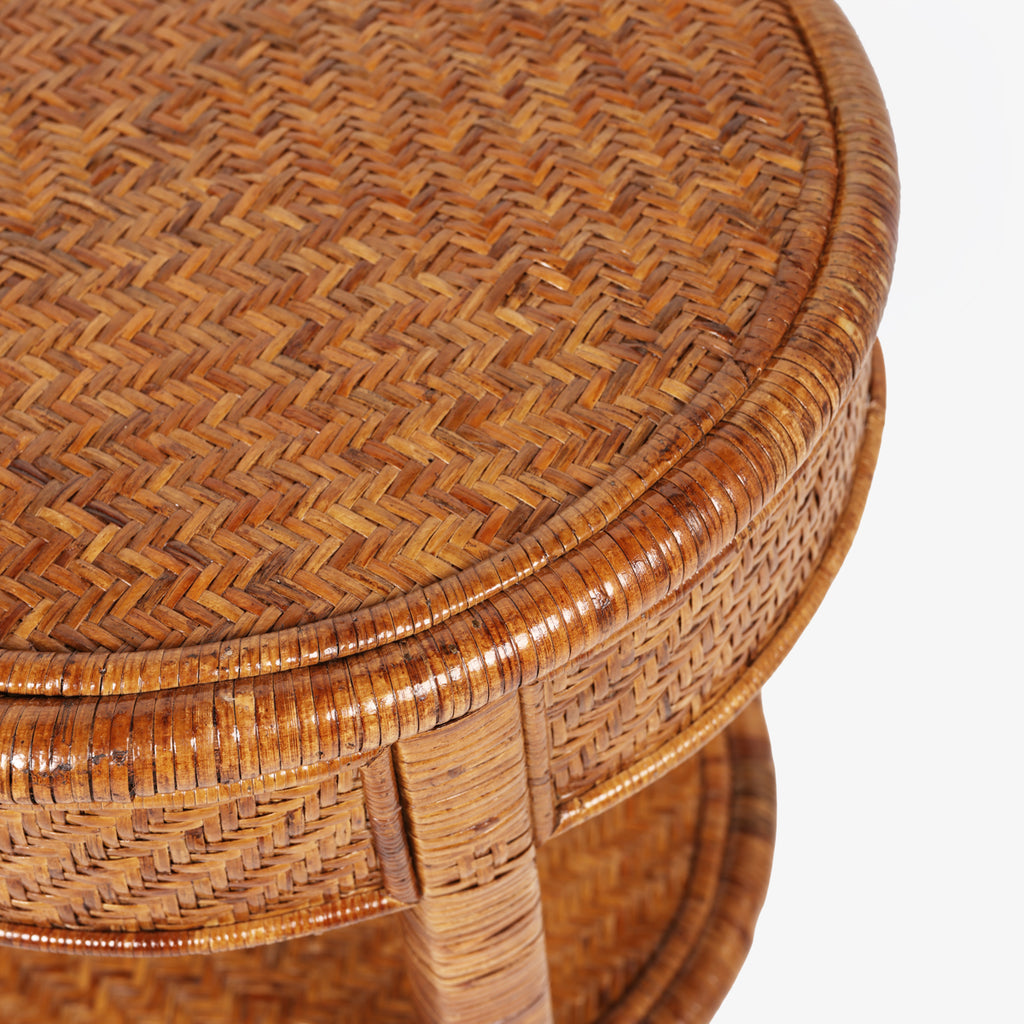 Close-up of a round woven rattan stool or side table showing the detailed herringbone pattern on the seat and backrest, with a warm brown finish against a white background.