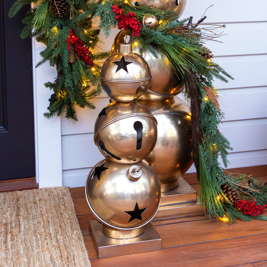 Stack of decorative gold bell ornament with star designs on a front porch with Christmas garland in the background.