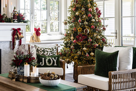 Traditional Christmas red and green baubles on a Christmas tree with green velvet Christmas cushions on brown rattan chairs in the foreground.