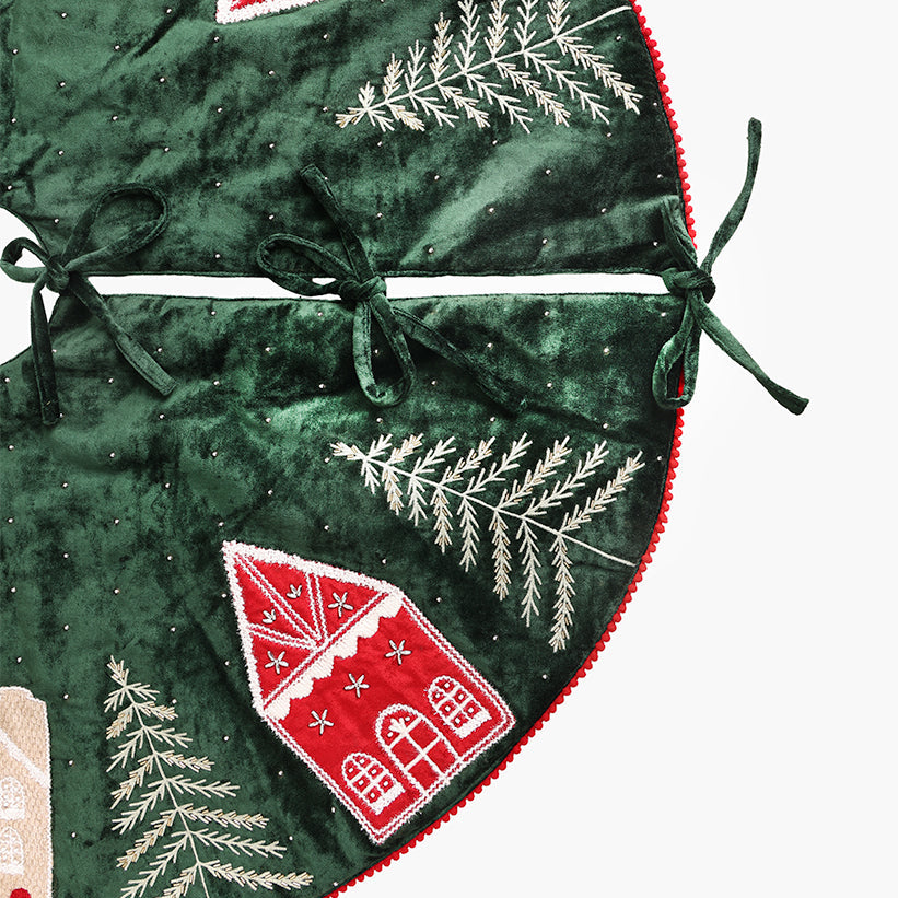 A close-up of a dark green velvet Christmas tree skirt with embroidered red gingerbread houses.